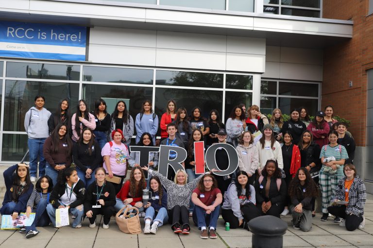 Two group photos of TRIO students standing outside campus buildings during program visits. In the left image, approximately 30 students pose together, with several holding large letters that spell "TRIO." In the right image, a larger group of approximately 50 students gathers in front of a building entrance, again posing with oversized "TRIO" letters. Students are smiling, standing, kneeling, and sitting together, highlighting participation, community, and engagement in the TRIO program.