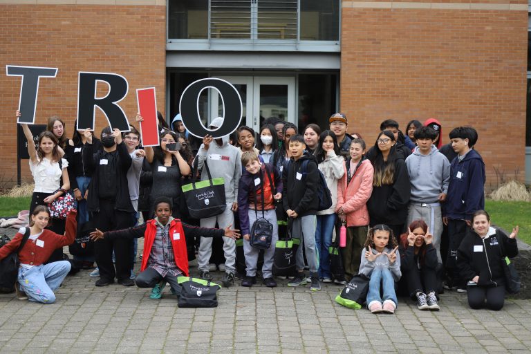 Two group photos of TRIO students standing outside campus buildings during program visits. In the left image, approximately 30 students pose together, with several holding large letters that spell "TRIO." In the right image, a larger group of approximately 50 students gathers in front of a building entrance, again posing with oversized "TRIO" letters. Students are smiling, standing, kneeling, and sitting together, highlighting participation, community, and engagement in the TRIO program.